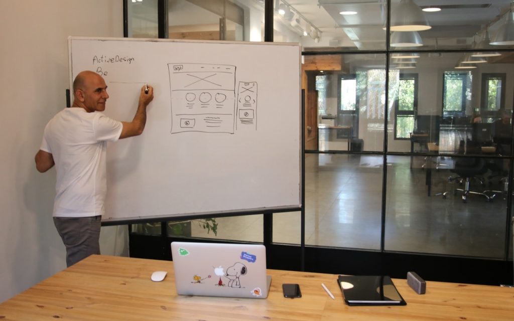 A man stands at a whiteboard drawing a website wireframe in a modern office. A wooden table with a laptop and a notebook is in the foreground. The office has glass walls and empty desks.