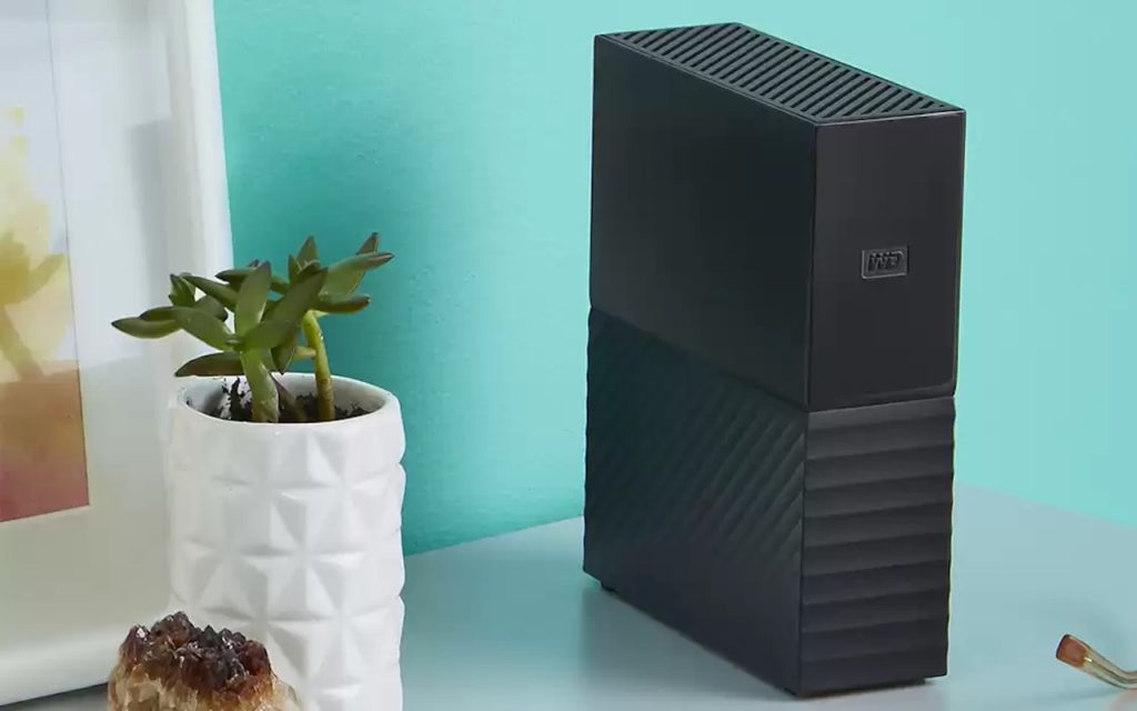 A black external hard drive sits on a desk next to a white geometric-patterned plant pot with a small succulent, against a light blue wall.