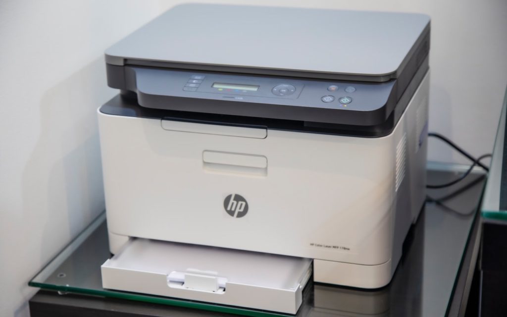 A white HP printer with scanning and copying functions sits on a glass surface against a white wall. Its paper tray is open, and control buttons and a small display screen are visible on the top panel.