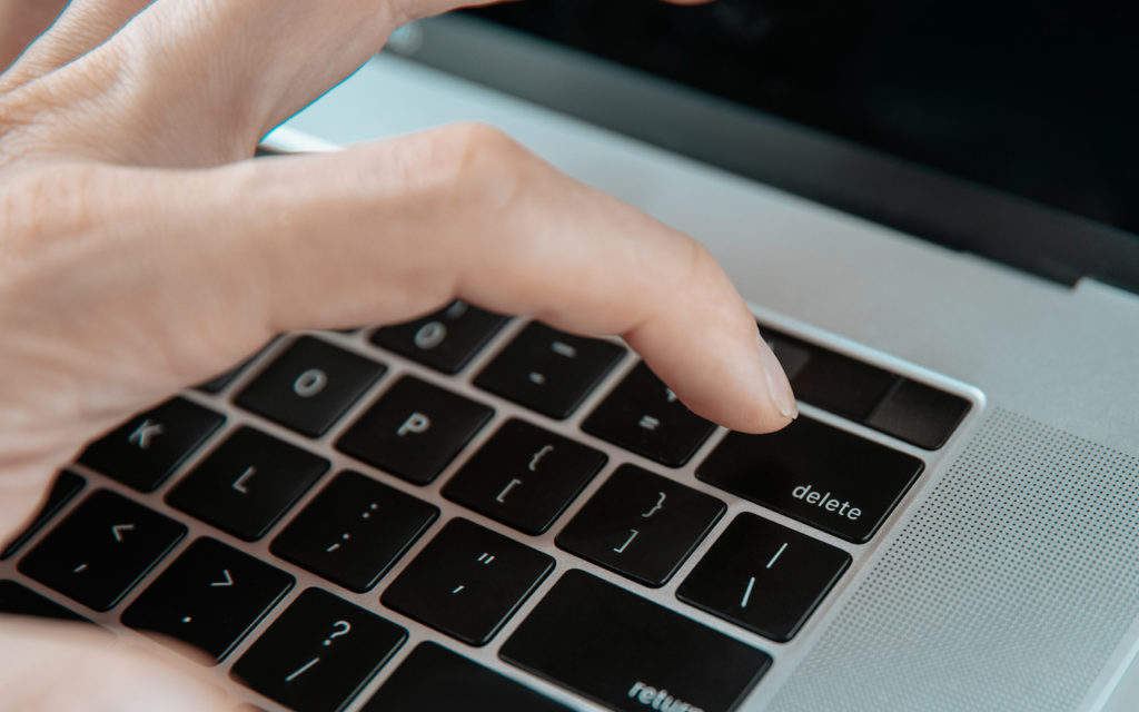 A close-up of a persons hand pressing the delete key on a laptop keyboard. The focus is on the finger and the keyboard, with the hand positioned above the black keys.