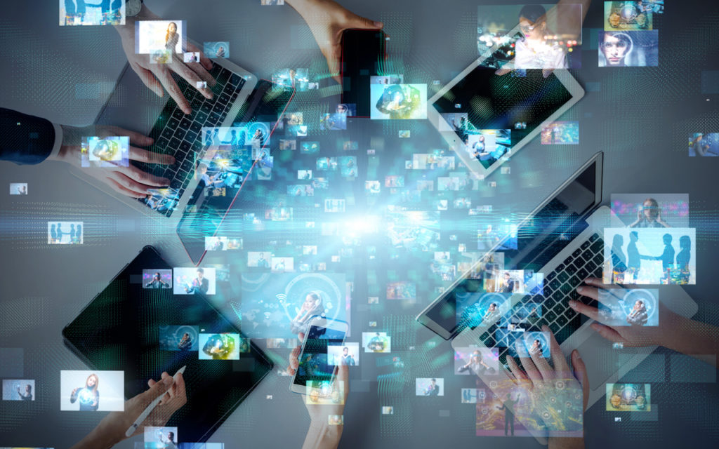 Several hands use laptops, tablets, and smartphones on a table, while digital images and holographic visuals float above, illustrating technology, communication, and multimedia connectivity.