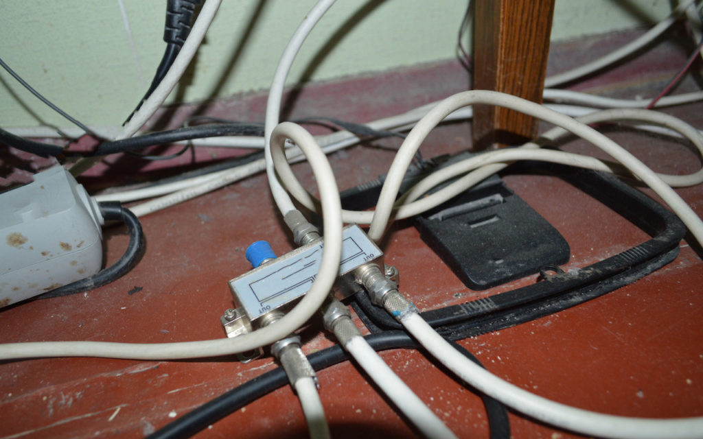 Several white and black cables are tangled on a red floor next to a wall. A splitter box connects some of the cables, and a wooden table leg and power strip are visible nearby.