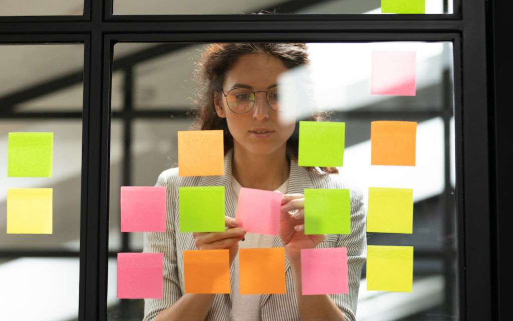 A woman stands behind a glass wall covered with colorful sticky notes. She is looking at and adjusting a pink note while wearing glasses and a striped blazer, in a modern office setting.