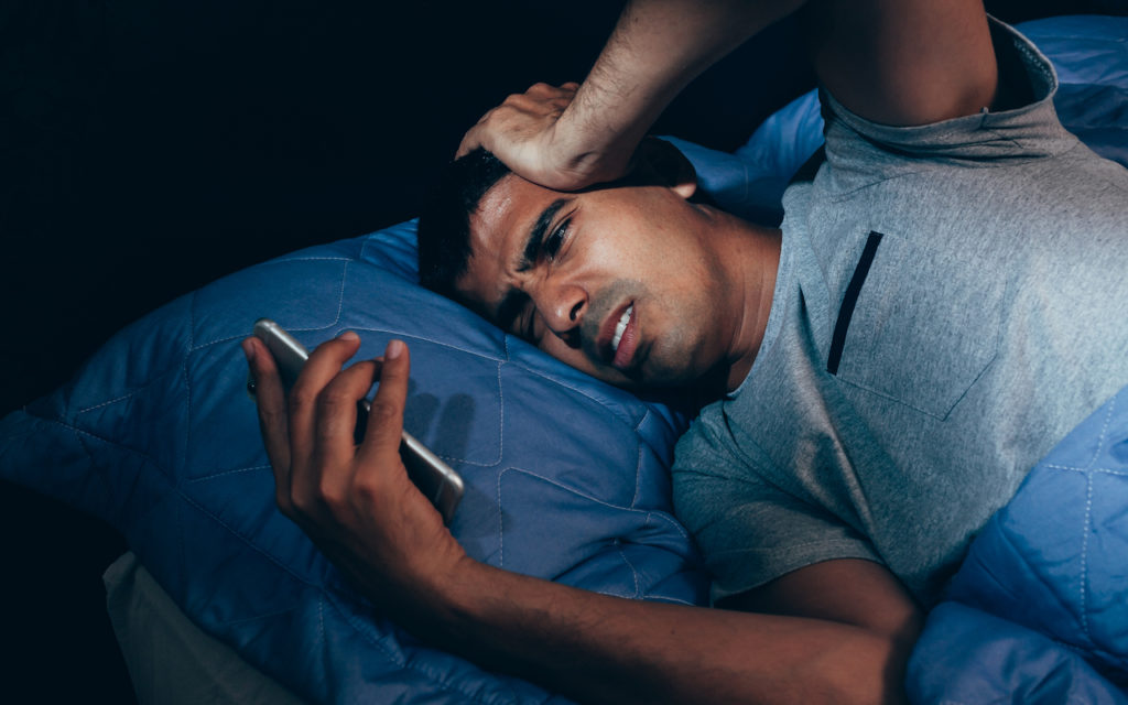 A man lying in bed under a blue blanket looks stressed and worried as he holds his smartphone, staring at the screen with a troubled expression.