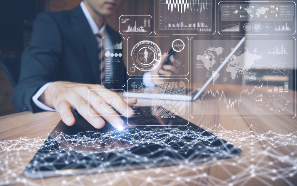 A person in a suit uses a tablet, with futuristic digital data graphs and infographics overlaid, while also holding a smartphone and sitting at a desk with a laptop.