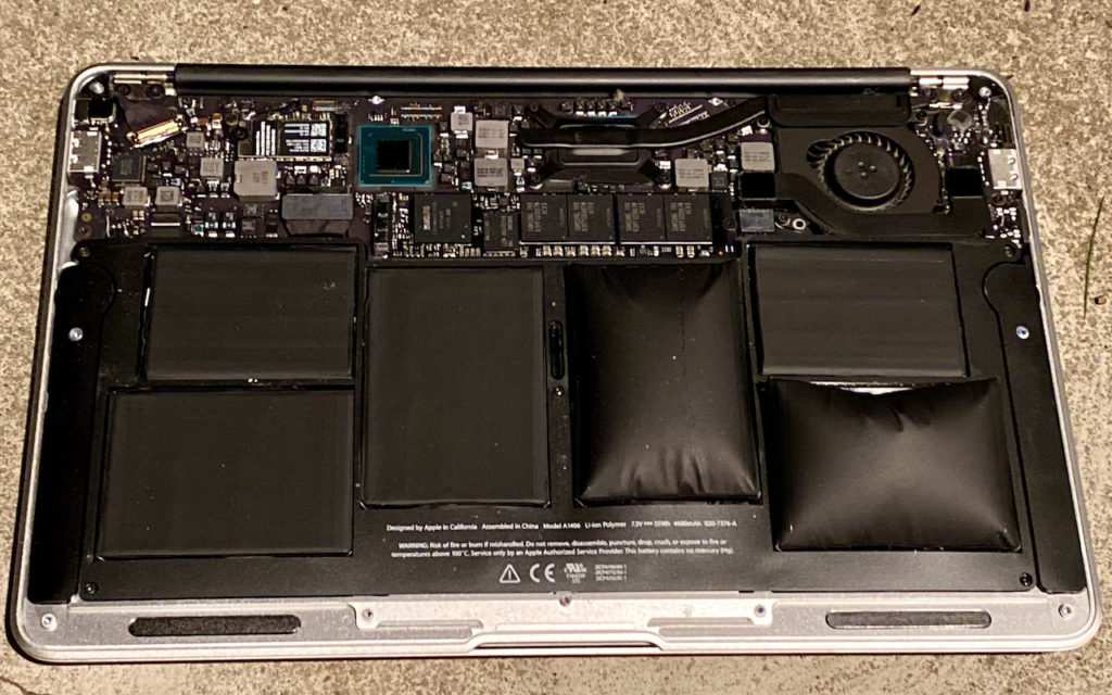 The inside of a laptop is shown with the back cover removed. Several battery cells are visibly swollen, with internal components, circuit boards, and a cooling fan surrounding them.