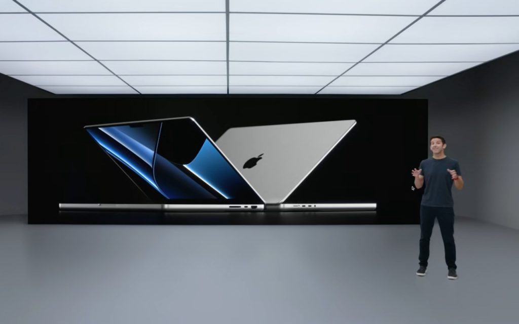 A presenter stands in a modern, minimalist room with a large screen behind him displaying two open MacBook laptops showcasing their design and Apple logo.