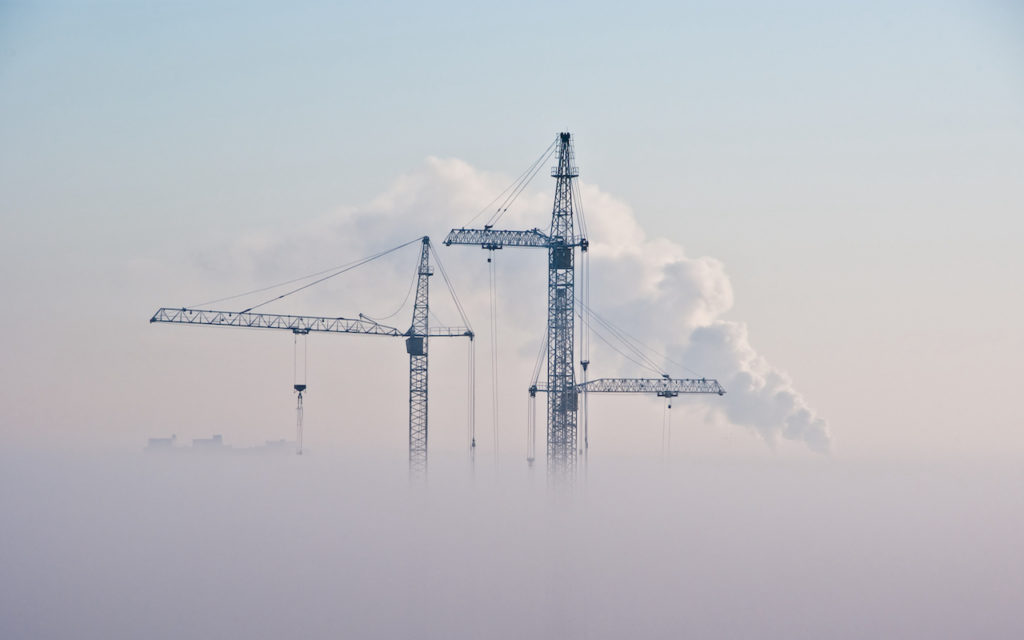 Three tall construction cranes rise above thick fog, with white smoke billowing from an industrial chimney in the background against a pale sky.
