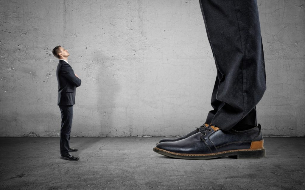 A small man in a suit stands facing a much larger man, whose legs and shoes dominate the foreground, against a gray concrete wall. The image emphasizes the size difference between the two figures.