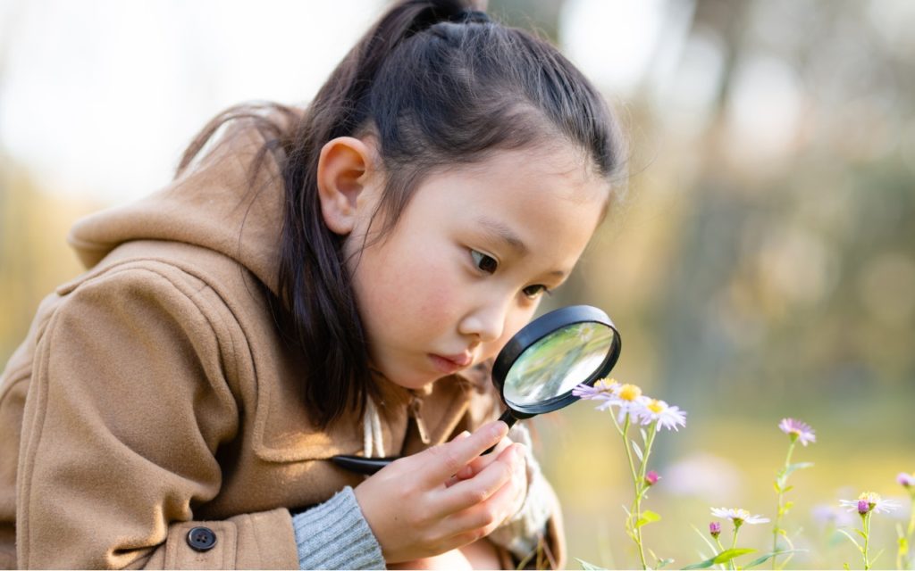 A young girl wearing a brown coat closely examines small flowers outdoors using a magnifying glass on a sunny day.