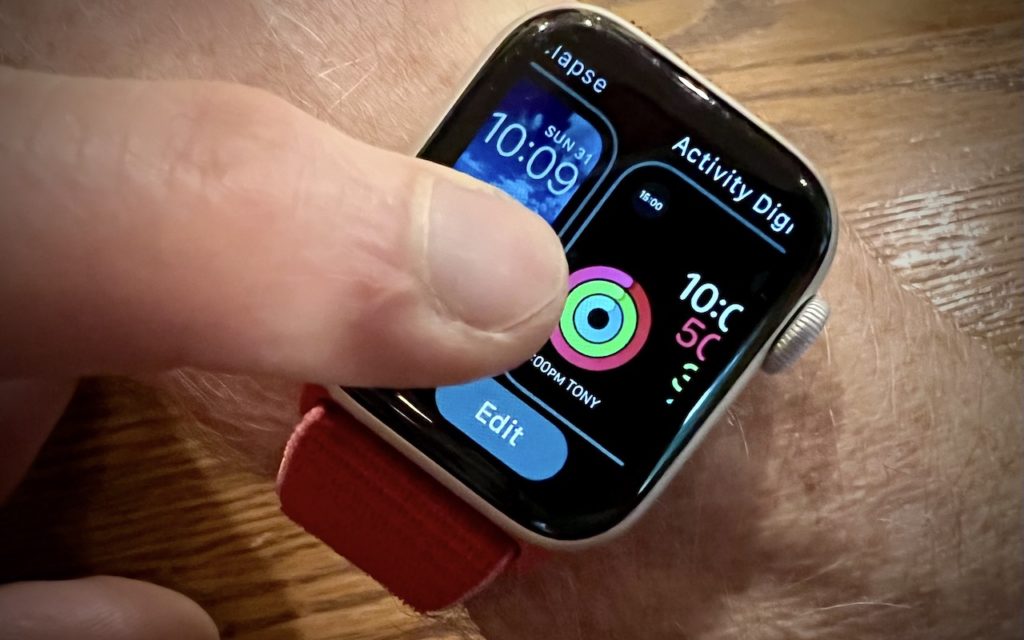A close-up of a person’s wrist wearing a smartwatch with a red band. The hand is touching the watch screen, which displays time, weather, and activity rings. The wooden surface beneath the wrist is visible.