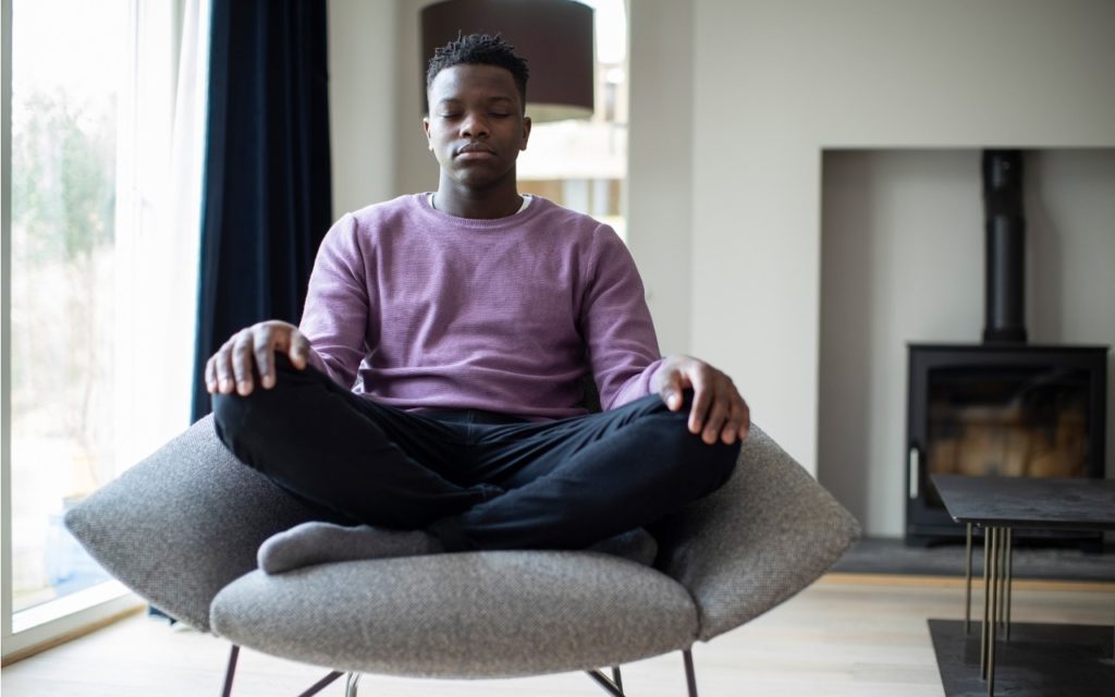 A person sits cross-legged on a modern chair indoors, eyes closed and hands resting on their knees, appearing calm and meditative. The setting is a bright room with a fireplace and large windows.
