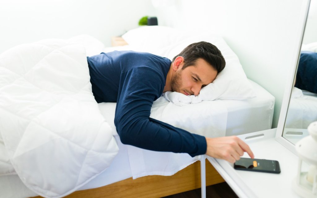 A man lies in bed under a white comforter, reaching out to turn off the alarm on his smartphone on a bedside table, looking tired and sleepy.