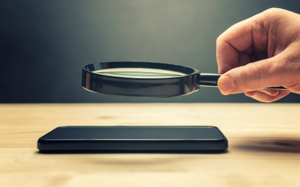 A hand holds a magnifying glass over a smartphone lying on a wooden surface, with a dark background.