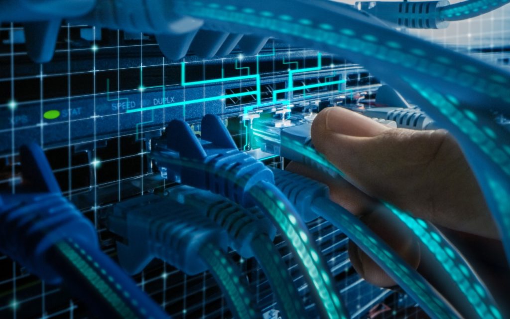 A hand connects a blue network cable to a server with multiple Ethernet cables and ports, overlaid with digital grid and glowing technology effects, representing data connections and internet infrastructure.