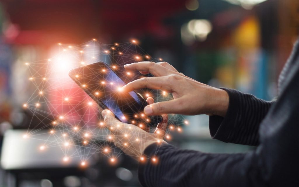 A person uses a smartphone with one hand while touching the screen with the other. Abstract glowing network connections are superimposed, symbolizing digital connectivity and communication.