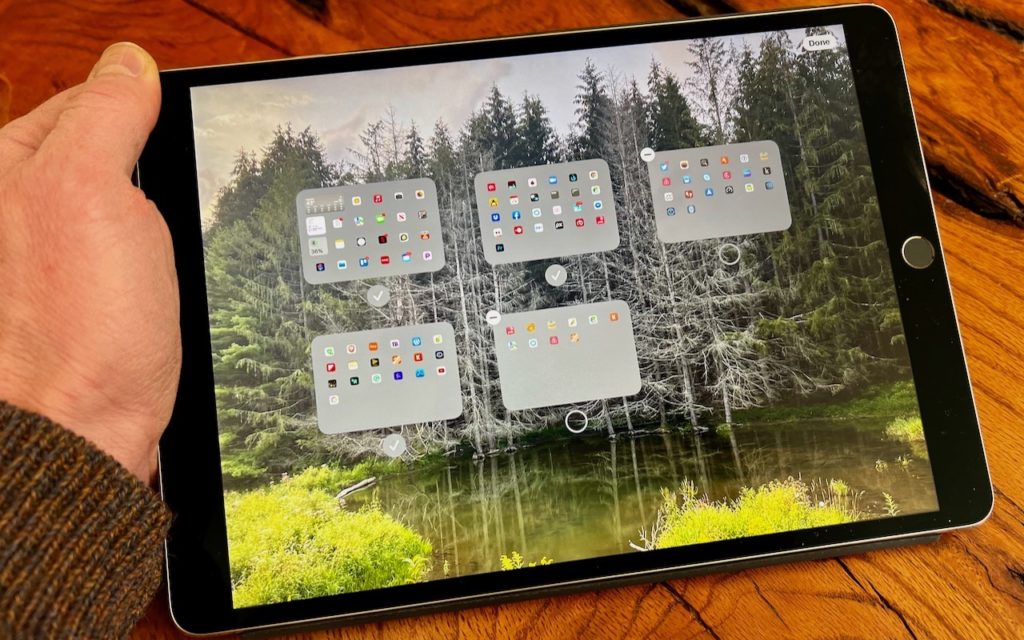A hand holding a tablet with an app-filled home screen displayed over a background image of a green forest and a small pond. The tablet has five app folders visible in the center of the screen.