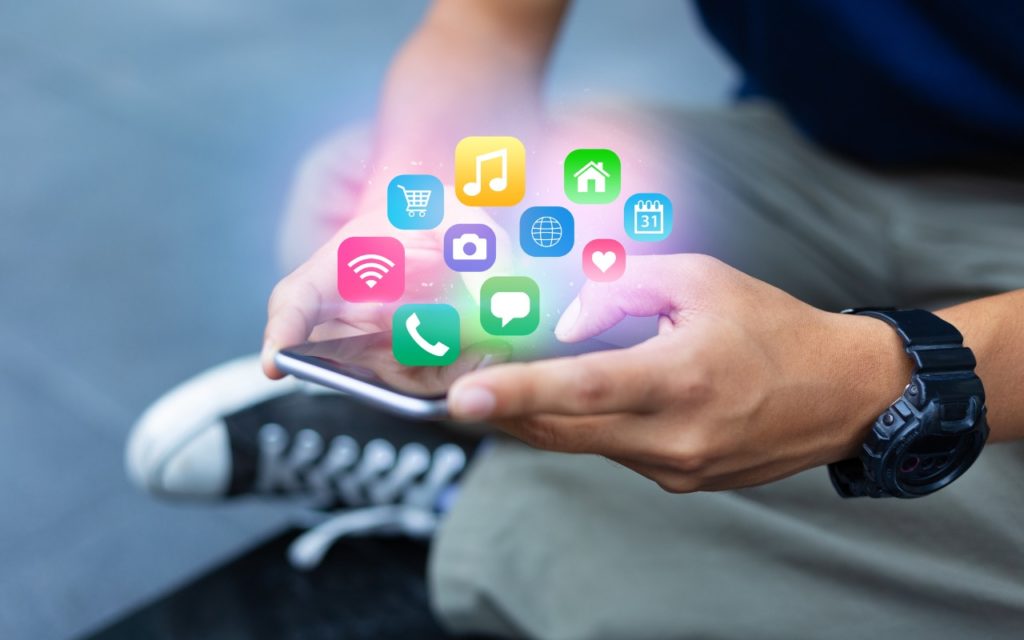 A person holds a smartphone with colorful app icons, such as music, shopping, calendar, and camera, appearing above the device. The person is wearing a dark shirt and a black watch.