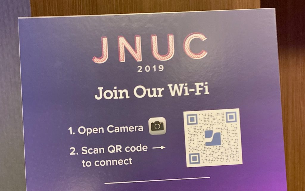 A sign reads JNUC 2019 Join Our Wi-Fi with instructions: 1. Open Camera 2. Scan QR code to connect. There is a QR code and a camera icon on a purple background.