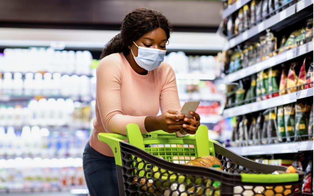 A woman wearing a protective face mask pushes a shopping cart filled with groceries while looking at her smartphone in a supermarket aisle. Shelves stocked with various food items are visible beside her.