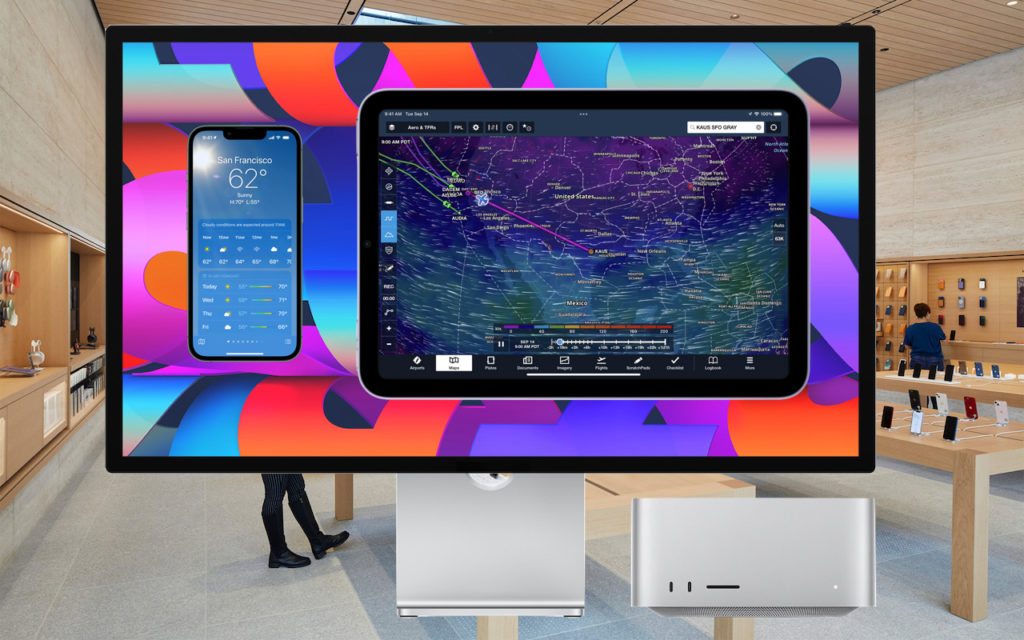 A large Apple monitor in a retail store displays a weather app and a flight tracking map, with colorful abstract wallpaper in the background. A Mac Studio and accessories are visible on the desk below.