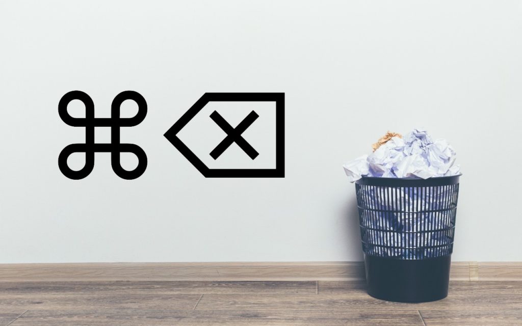 A black trash bin filled with crumpled paper sits on a wooden floor against a white wall. On the wall are large symbols for “Command” and “Delete.”.