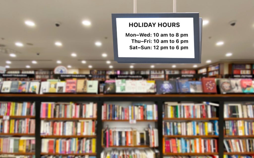 A sign hanging in a bookstore displays holiday hours: Mon–Wed 10 am to 8 pm, Thu–Fri 10 am to 6 pm, and Sat–Sun 12 pm to 6 pm. Shelves filled with books are visible in the background.