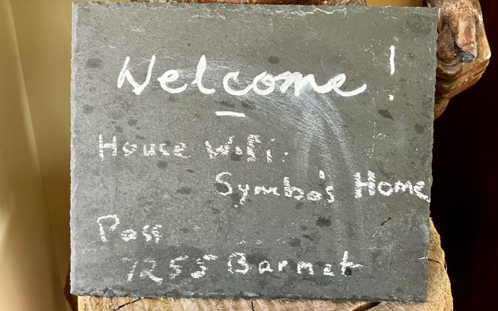 A small chalkboard sign with handwritten text: Welcome! House WiFi: Symbas Home. Pass: 1255Barnet. The board is resting on a wooden surface.