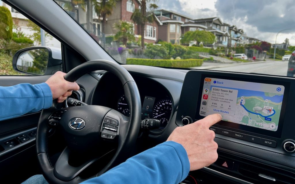 A person driving a Hyundai car uses the touchscreen navigation system, with a neighborhood of houses and palm trees visible through the windshield on a cloudy day.