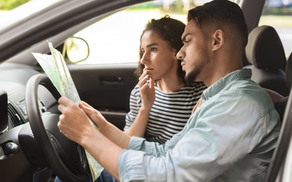 A man and woman sitting in a car look intently at a map, appearing thoughtful and focused as they try to find directions. The woman has her hand near her mouth, and both seem concentrated on the map.