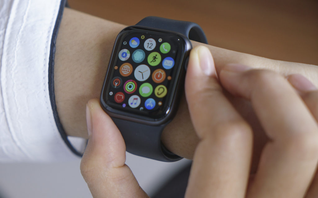 A person wearing a black smartwatch with a colorful app grid on the screen is touching the side button with their right hand. The person is dressed in a white long-sleeve shirt.