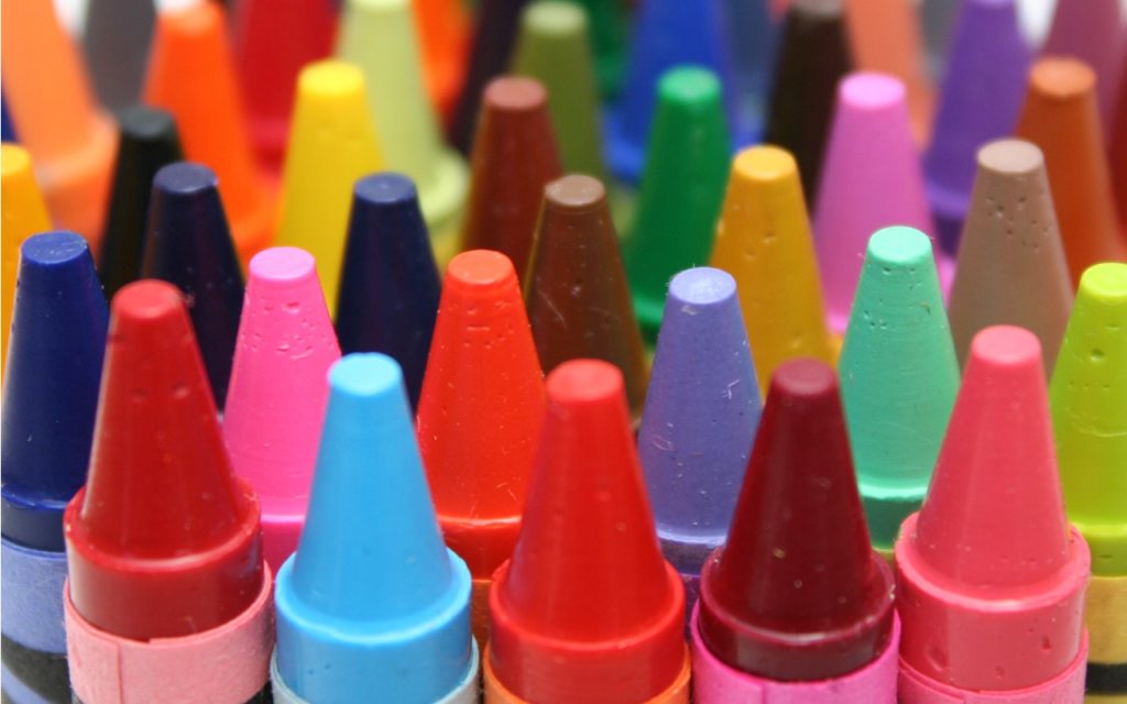 Close-up of the tips of many colorful crayons arranged closely together in rows, showing a variety of bright colors including red, blue, green, yellow, orange, pink, purple, and more.