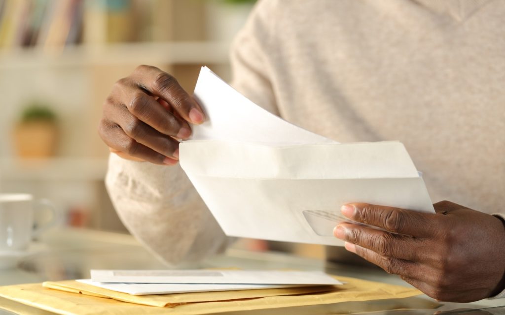 A person’s hands opening a white envelope, with other envelopes and papers on a table. The background is softly blurred and the person is wearing a light sweater.