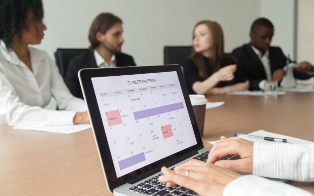 A person types on a laptop displaying a digital calendar while four businesspeople sit at a conference table in a meeting or discussion.