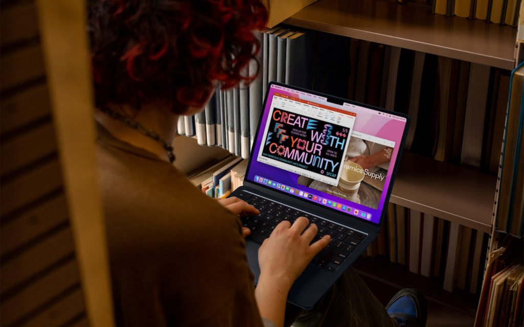 A person with curly hair types on a laptop, sitting between bookshelves. The laptop screen displays a colorful presentation slide reading Create with your community alongside images and text.