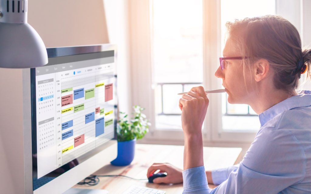 A person wearing glasses sits at a desk, looking thoughtfully at a large computer monitor displaying a colorful digital calendar or schedule. A potted plant and desk lamp are visible nearby.