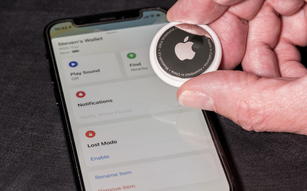 A hand holds an Apple AirTag above an iPhone displaying the Find My app, showing tracking options and settings for a device labeled Stevens Wallet.