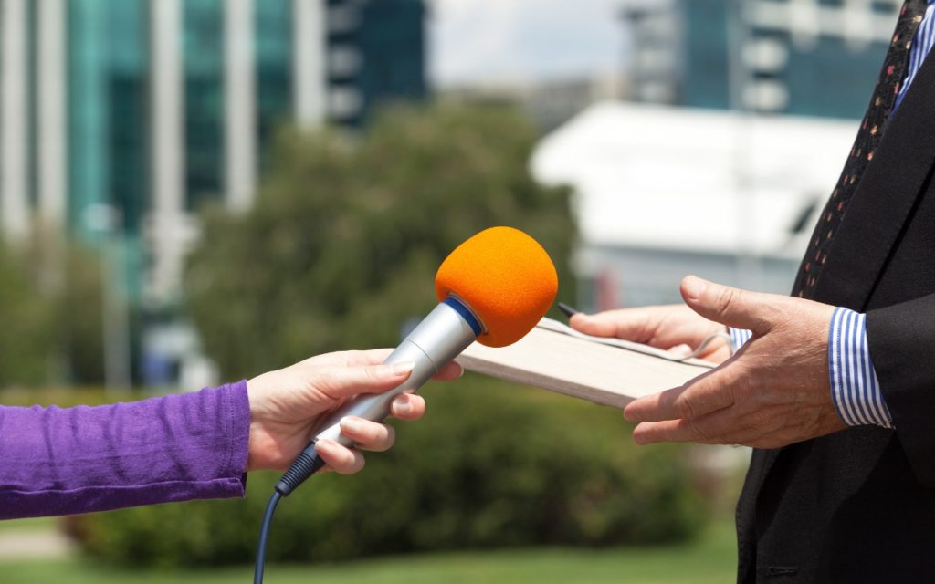 A person holds a microphone with an orange foam cover toward another person in a suit who is holding a book, conducting an outdoor interview with buildings blurred in the background.
