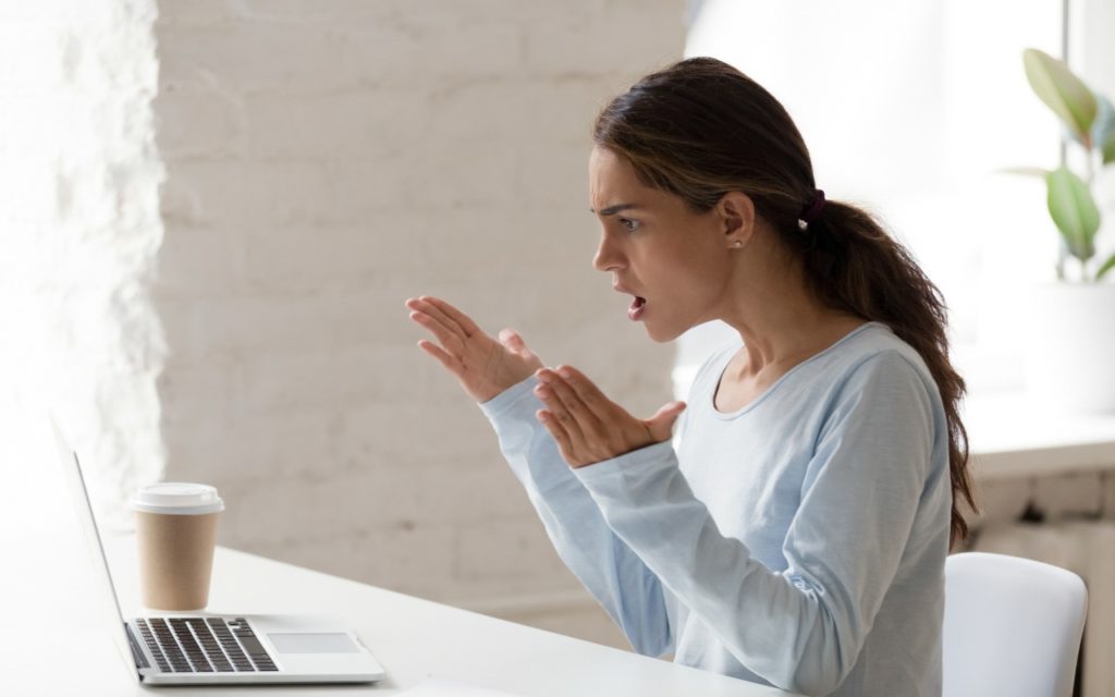 A woman sits at a desk in front of a laptop, looking frustrated or upset, with her hands raised in exasperation. A takeaway coffee cup is on the desk, and a plant is visible in the background.