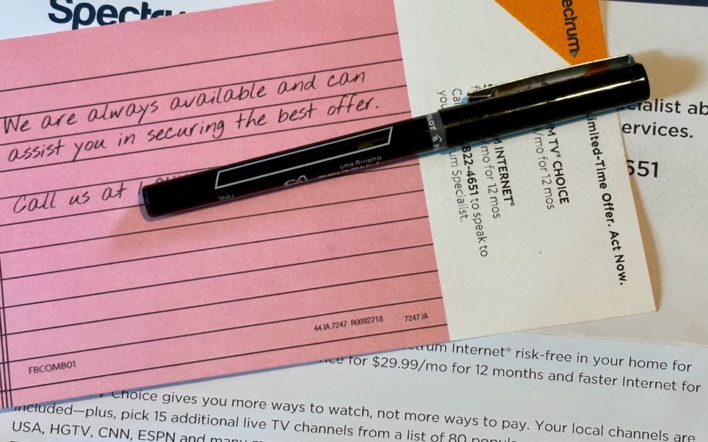 A black pen rests on a pink index card with handwritten text and two Spectrum mail advertisements. The card reads, “We are always available and can assist you in securing the best offer. Call us at …”.