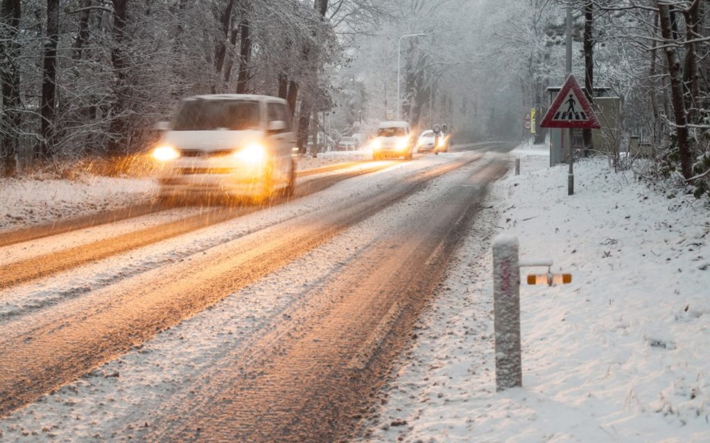 Cars drive carefully on a snow-covered road lined with trees, headlights on in low visibility. Snow blankets the ground and branches, and a traffic warning sign is visible on the right side of the road.