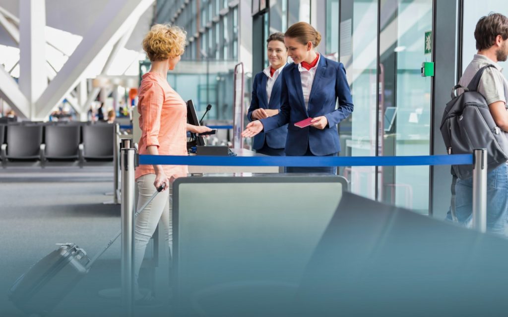 A female airline staff member in a blue uniform gestures for a woman with luggage to proceed at an airport boarding gate, while another staff member and a man with a backpack stand nearby.