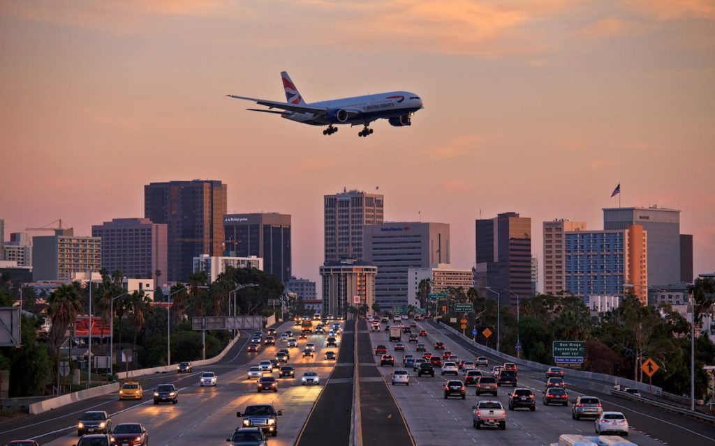 A British Airways airplane descends above a busy highway with cars during sunset, with a city skyline and tall buildings in the background.