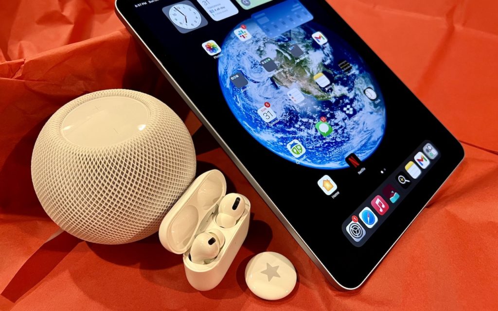 An iPad displaying a home screen with an Earth wallpaper sits on red paper, alongside a white HomePod Mini, AirPods Pro in an open case, and an Apple AirTag with a star sticker.