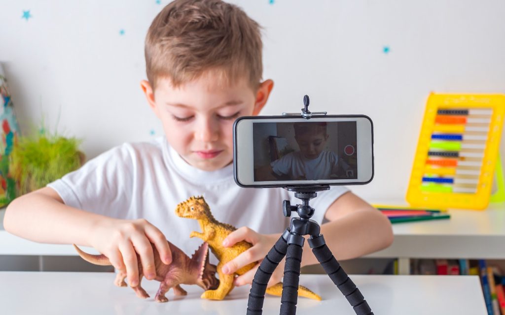 A young boy plays with two toy dinosaurs at a table while recording himself with a smartphone mounted on a tripod. A colorful xylophone and other toys are in the background.
