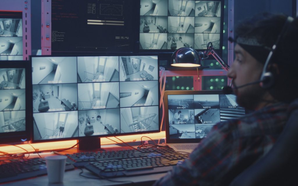 A person wearing a headset monitors multiple security camera feeds on several screens at a workstation in a control room. The screens display black-and-white footage of hallways and rooms.