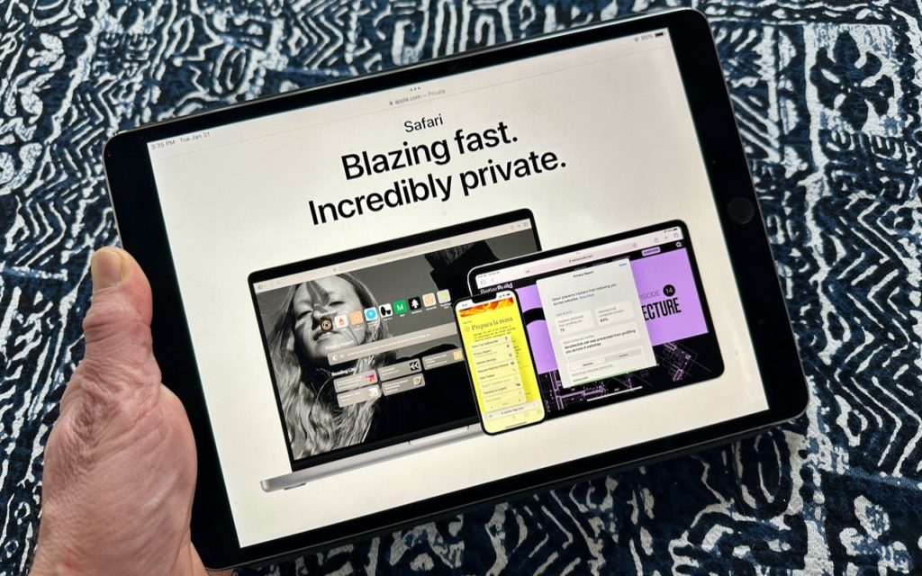 A hand holds an iPad displaying a Safari browser page with the text Blazing fast. Incredibly private. alongside images of a laptop and a tablet. The background is a blue and white patterned fabric.