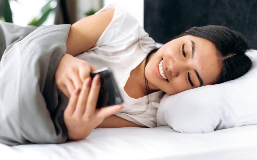 A young woman lies in bed under a gray blanket, smiling as she looks at her smartphone. She rests her head on a white pillow and appears relaxed and happy.