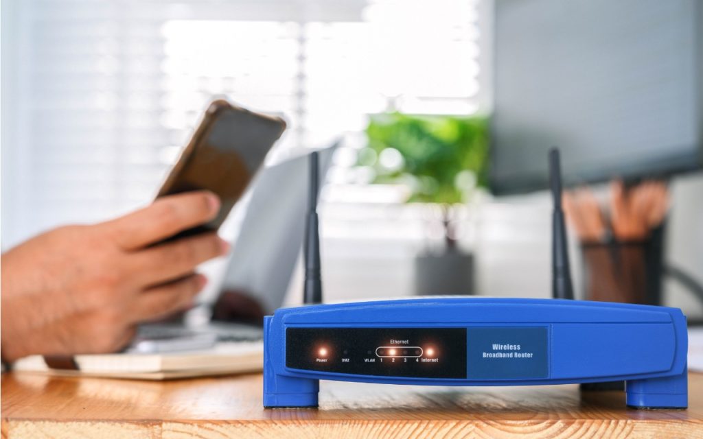 A blue wireless broadband router with indicator lights is on a wooden desk. In the blurred background, a person is using a smartphone and a laptop.