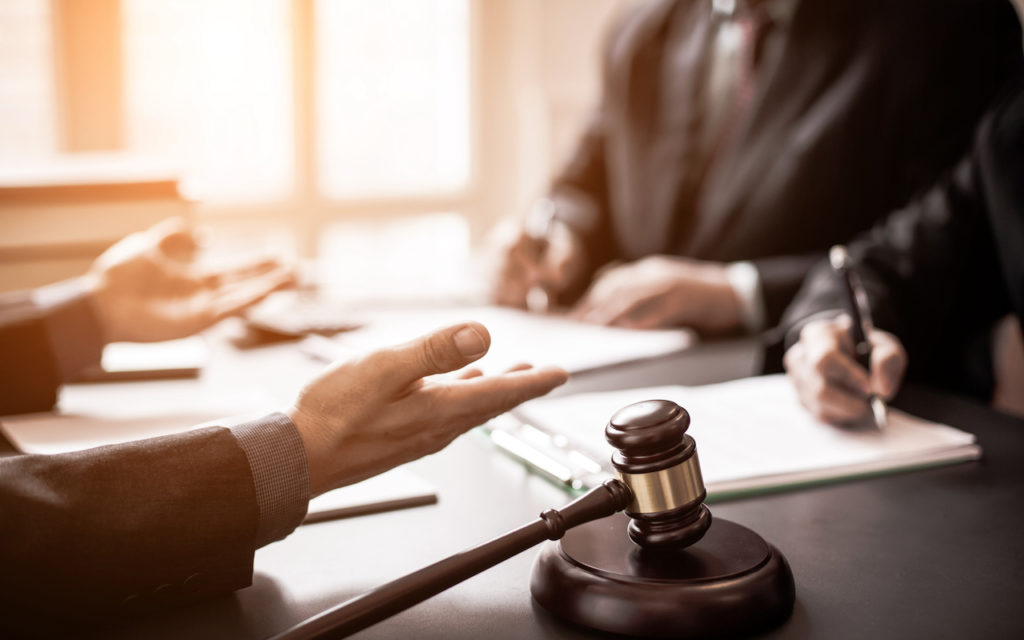 Two people sit at a desk with documents, one gesturing with open hands, the other writing. A wooden gavel rests on the table, suggesting a legal or courtroom setting.